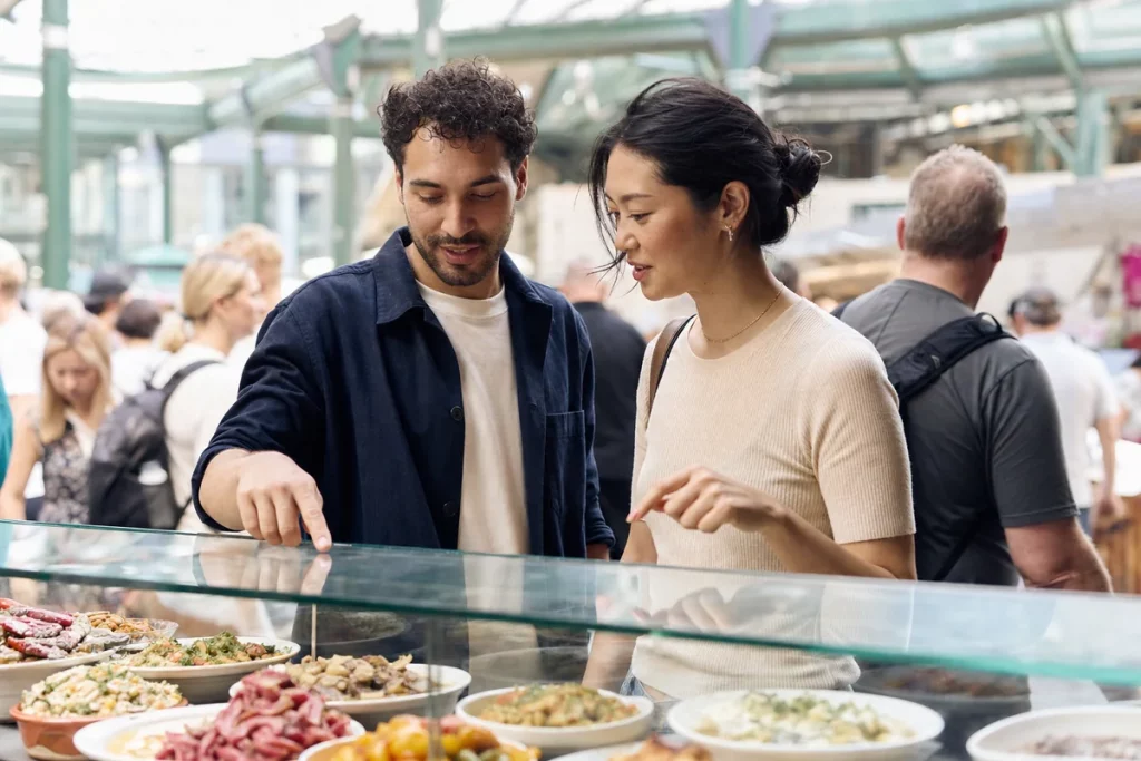 Two adults meeting for a second date at a lively public food market, choosing a small dish together while keeping the mood relaxed.