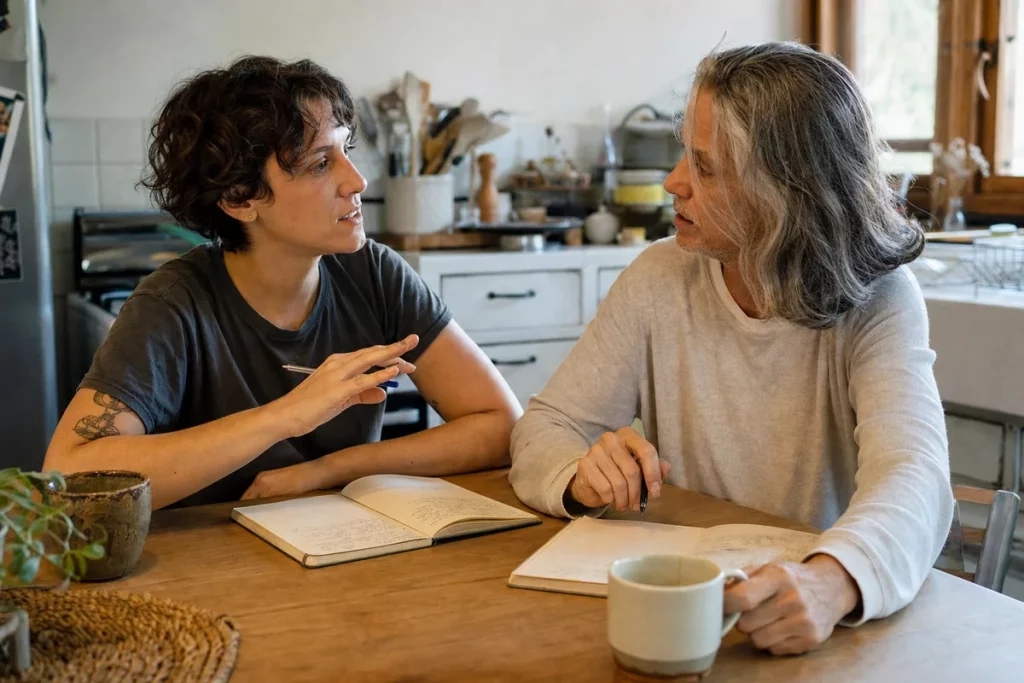 Two adult partners of visibly different ages talking as equals across a kitchen table, each with an open notebook and relaxed posture.